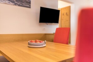 Une table à manger avec un saladier de fruits, un téléviseur fixé au mur et une chaise rouge.