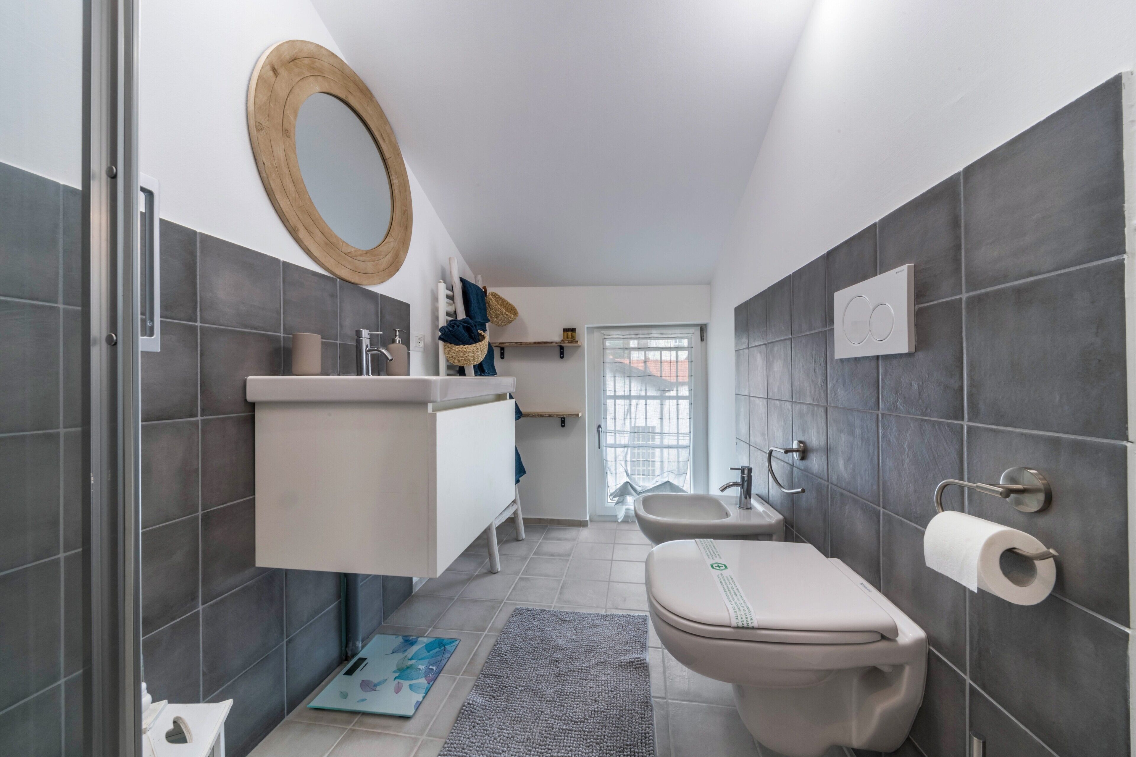 Un baño moderno con ducha, inodoro y lavamanos, con azulejos grises y un espejo con marco de madera.