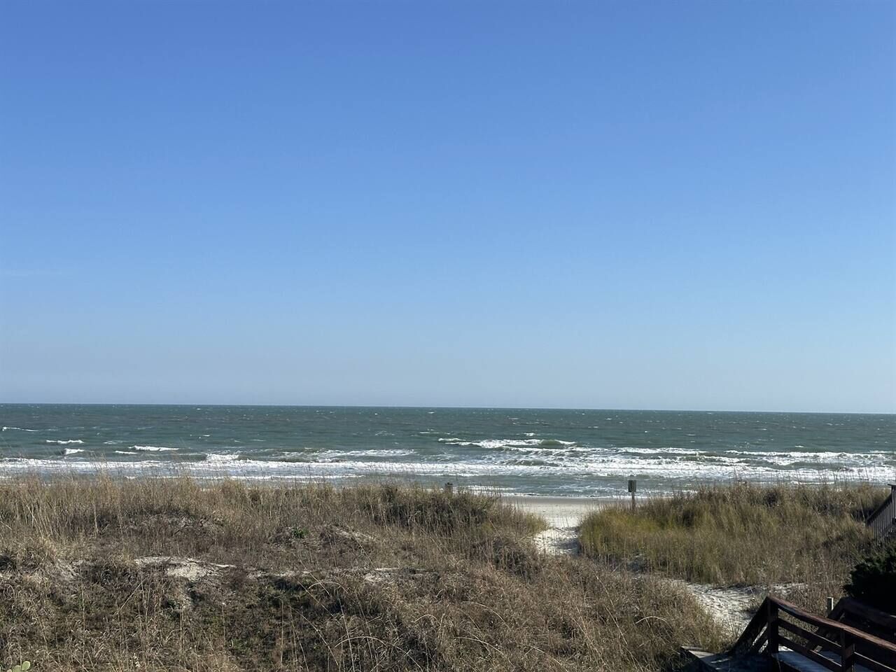 A beach with a wooden boardwalk leading to the water, a clear sky, and waves crashing onto the shore.
