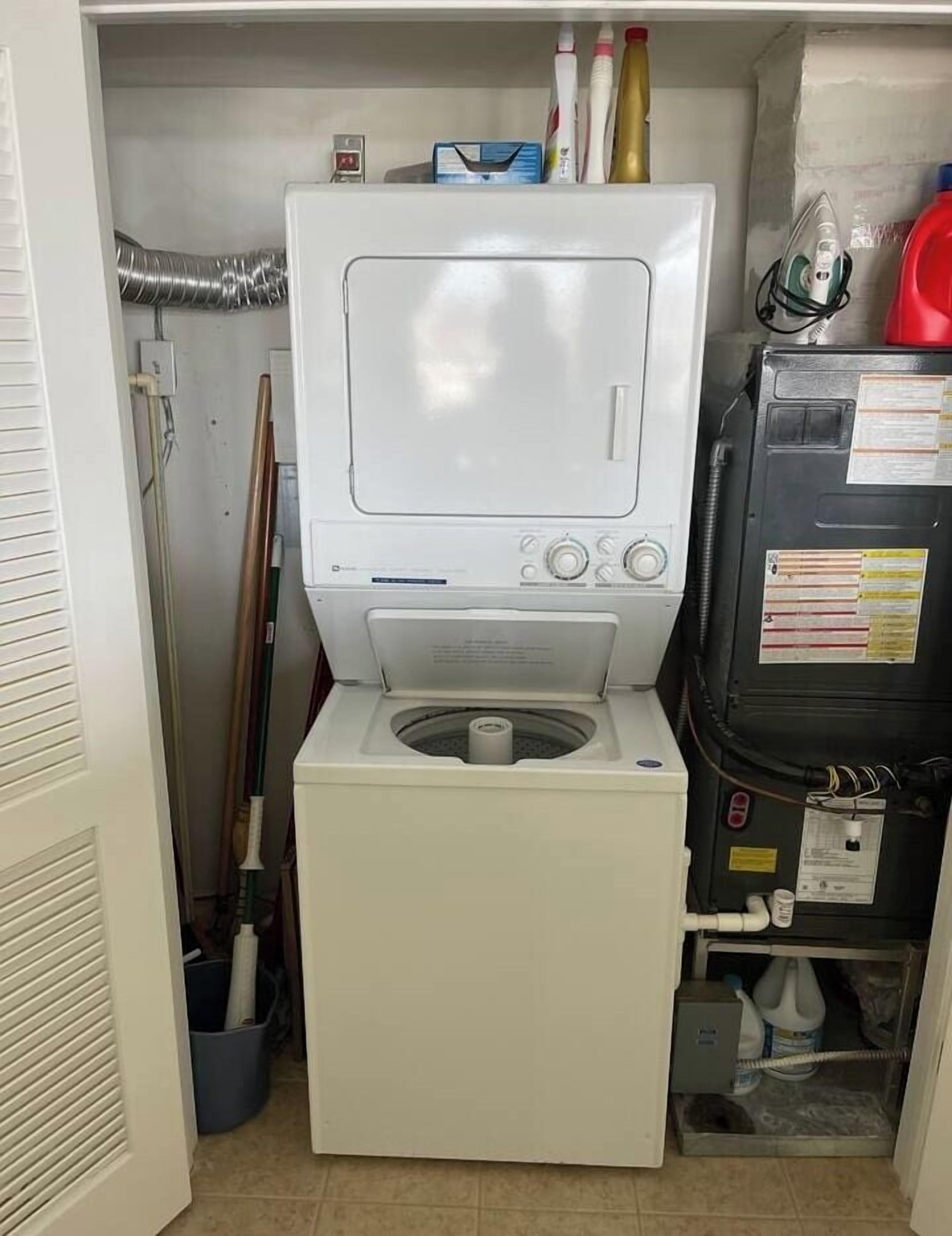 A compact laundry room with a washing machine and dryer, a metal vent, and cleaning supplies on the shelves.