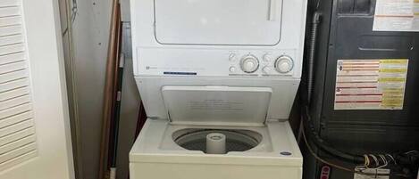 A compact laundry room with a washing machine and dryer, a metal vent, and cleaning supplies on the shelves.