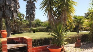 An outdoor brick barbecue grill with a stone wall, surrounded by a garden with palm trees and a lawn.
