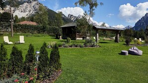 En rofylld trädgård med en lusthus, solstolar och en liten byggnad, omgiven av berg.