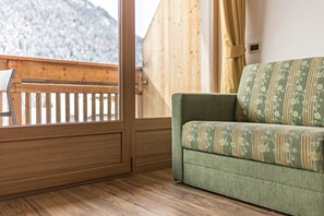 Une pièce dotée d’un plancher en bois, d’un canapé vert à motifs floraux, d’une rambarde en bois et offrant une vue sur une montagne enneigée.