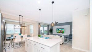 A modern kitchen with a central island, pendant lights, and a dining area with white chairs and a table.