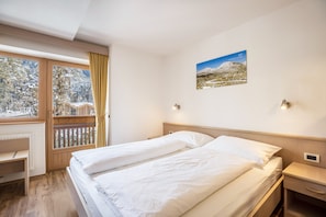 A hotel room with a bed, a desk, a chair, a window with a view of trees, and a wall-mounted picture. - Runcac - Holiday Apartment Salt with Mountain View, Wi-Fi & Balcony (San Vigilio)