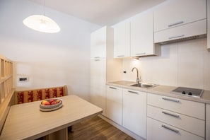 A modern kitchen with white cabinets, a stainless steel sink, and a wooden dining table with a bowl of fruit. - Runcac - Holiday Apartment Salt with Mountain View, Wi-Fi & Balcony (San Vigilio)