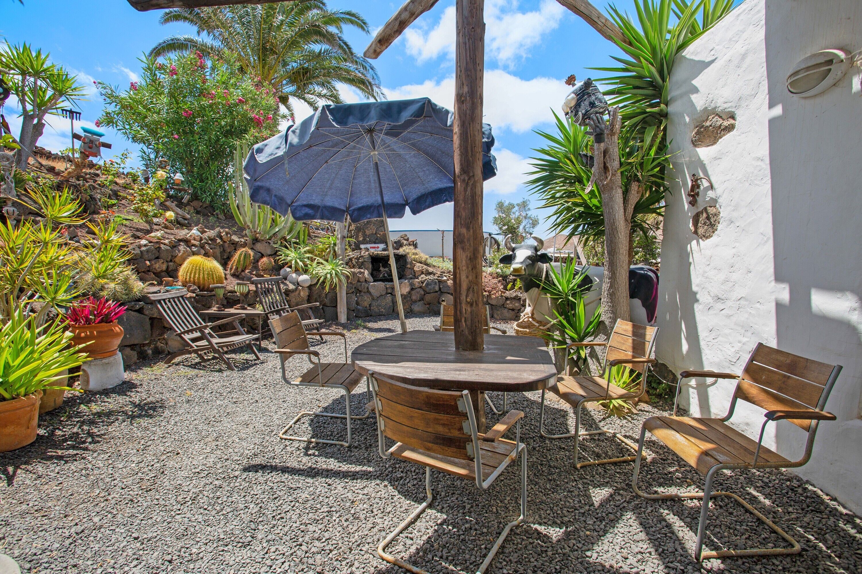Un patio al aire libre con una mesa redonda de madera, sillas de metal, un parasol azul y varias macetas con plantas.