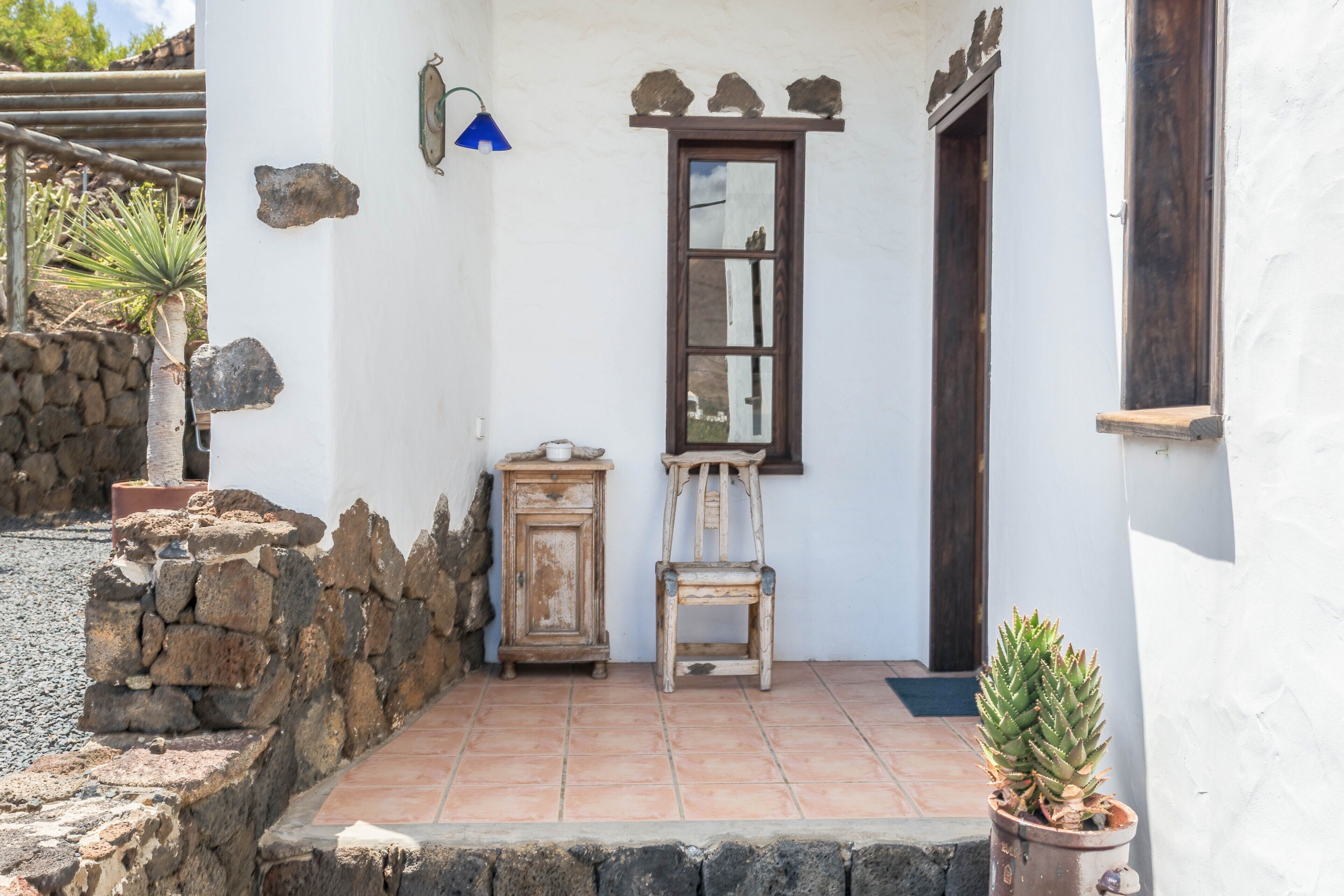 A white building with a stone wall, a wooden chair, a small cabinet, and a potted plant.