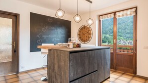 A kitchen with a central island, a chalkboard with writing, a round wooden clock, and a view of a forest through a window.