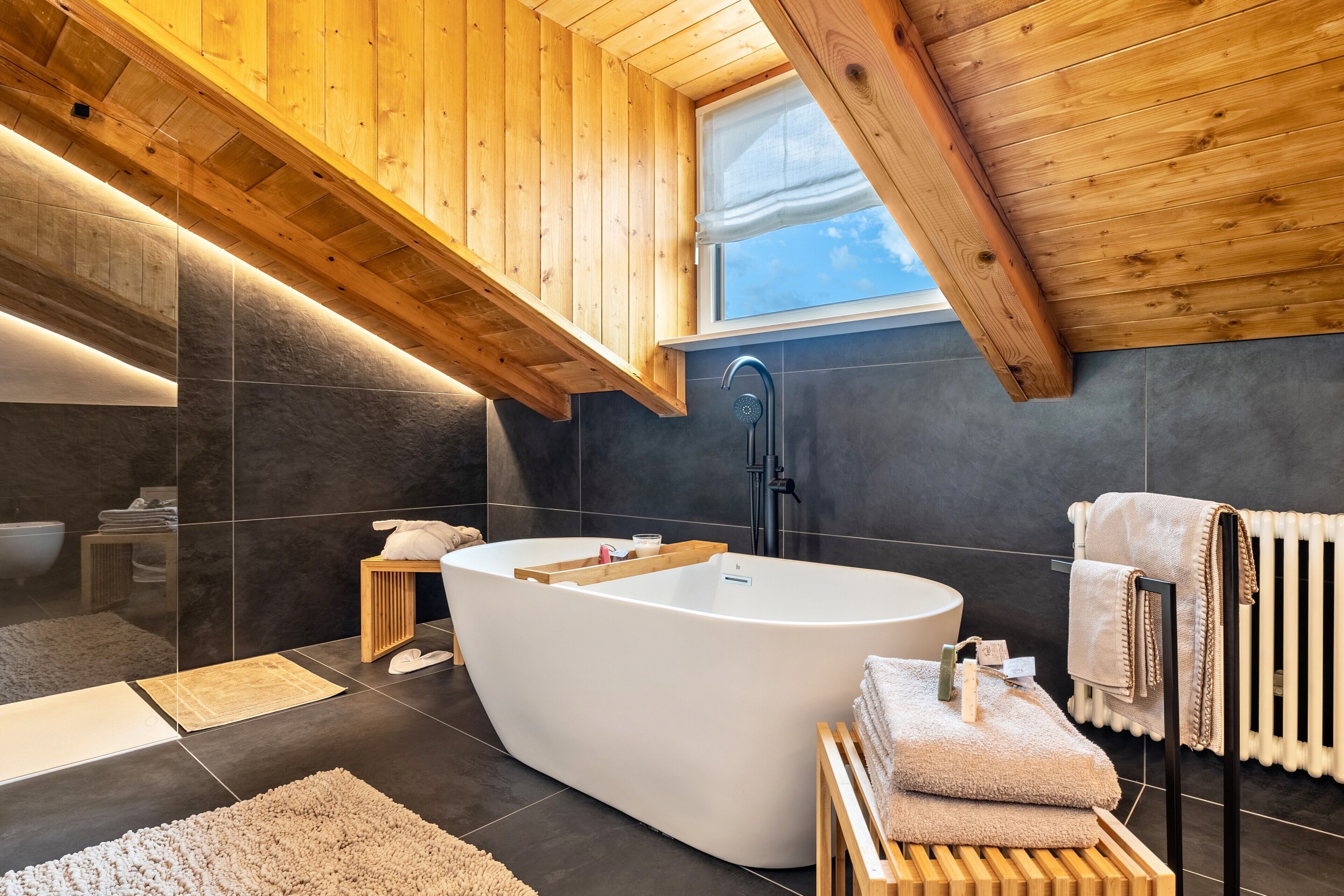 A modern bathroom with a freestanding bathtub, wooden ceiling beams, and a window.