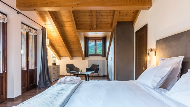 A bedroom with a large bed, wooden ceiling beams, a small table with chairs, and a window with curtains.
