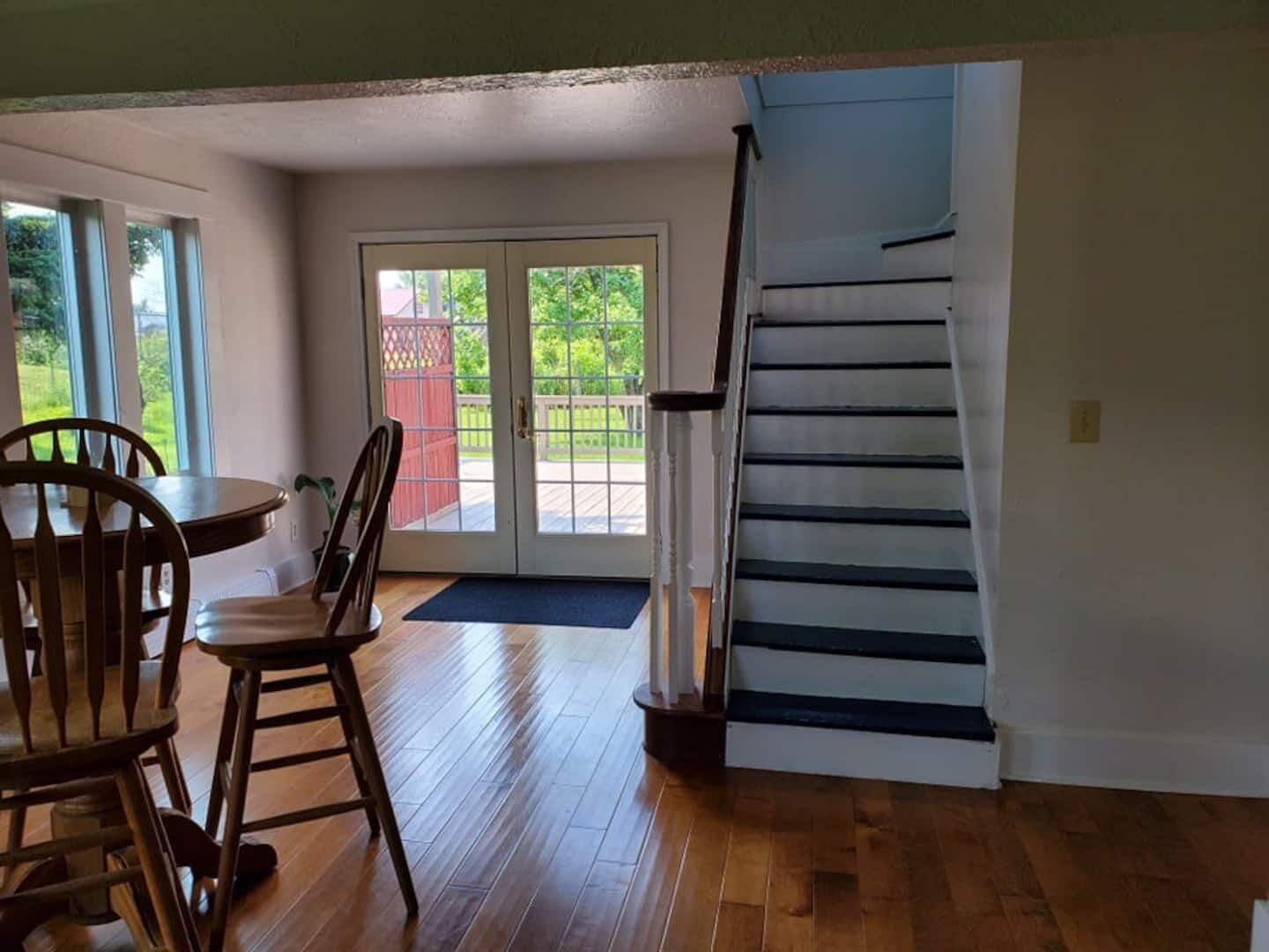 A spacious room with a wooden floor, a dining area with chairs and a table, a staircase, and a sliding glass door leading to an outdoor area.