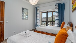 Ein Hotelzimmer mit zwei Betten, einem großen Fenster mit Vorhängen, einer Pendelleuchte und einem Spiegel an der Wand.