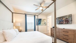 Une chambre à coucher avec un lit à baldaquin, un ventilateur de plafond, une table de chevet avec une lampe, une commode et une grande œuvre d’art encadrée au mur.