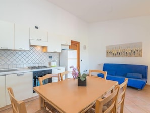 Une cuisine compacte avec des meubles blancs, une cuisinière et un évier. On y trouve une table à manger avec des chaises, ainsi qu’un canapé bleu dans l’espace salon.