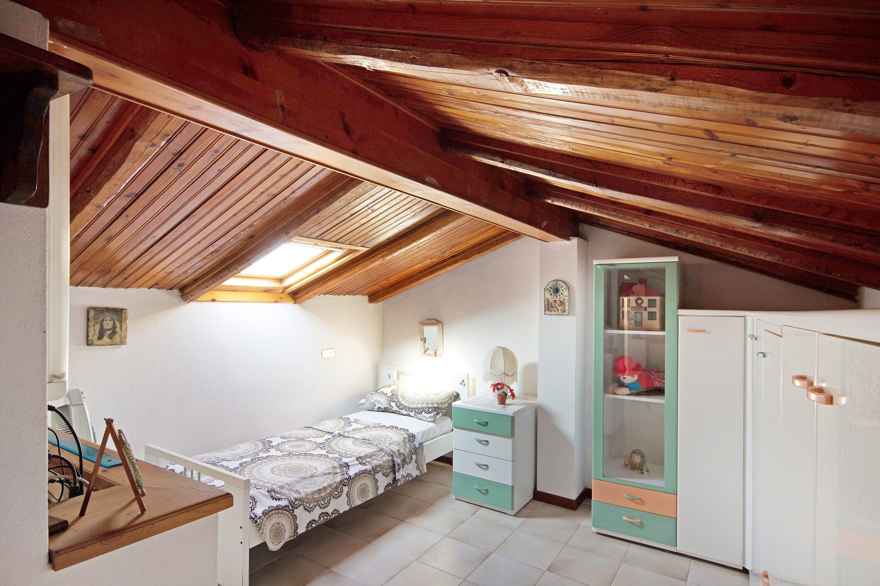 Ein Schlafzimmer mit schrägen Wänden, einem Bett, einem Schminktisch, einem Kleiderschrank und einem Schreibtisch mit Stuhl.