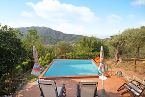 Una piscina rectangular con vistas a montañas al fondo, rodeada por un patio con sillas de descanso y sombrillas.