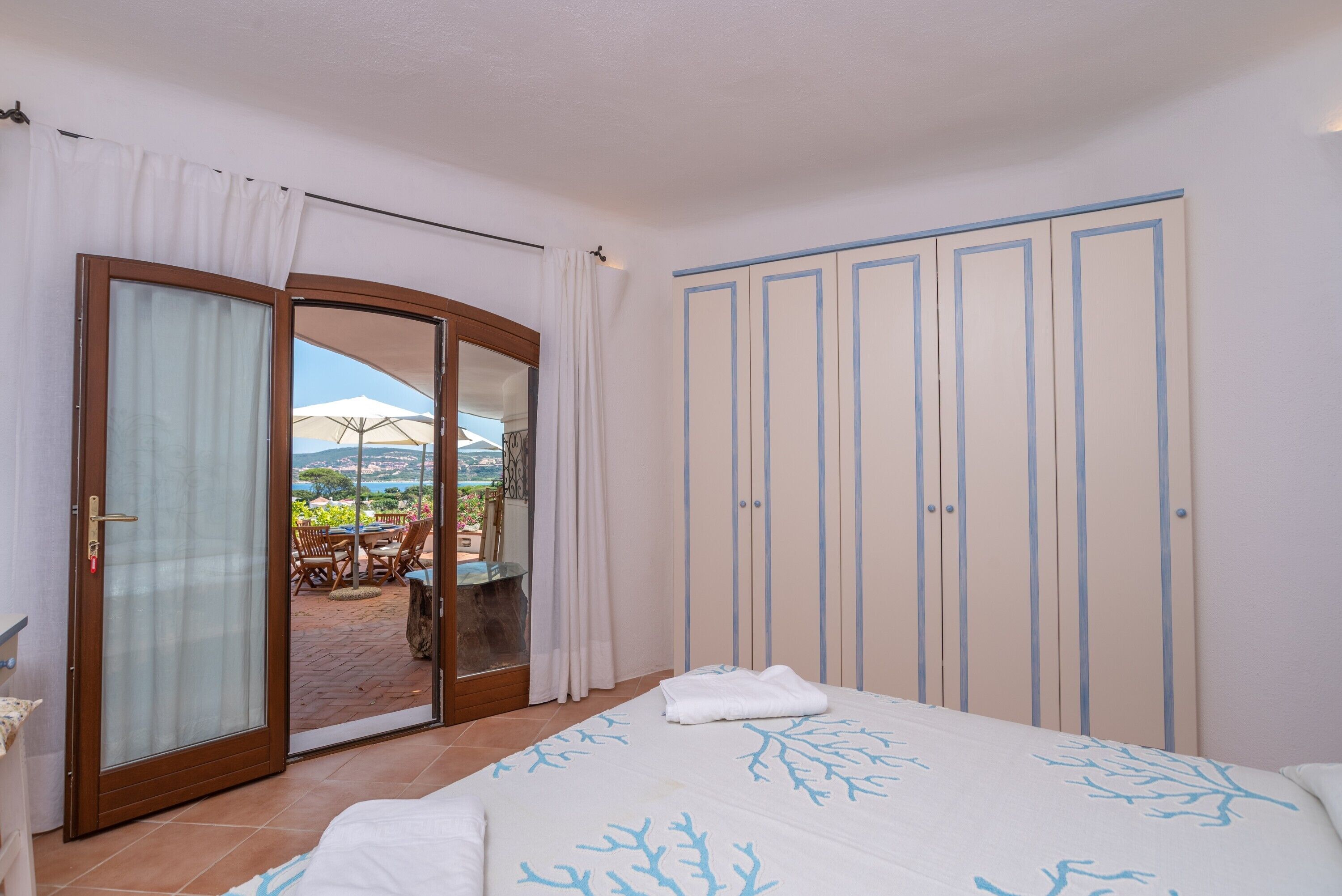 Ein Schlafzimmer mit einem Bett, einer Glastür zum Außenbereich mit Tisch und Stühlen sowie einem Kleiderschrank mit vertikalen Linien.
