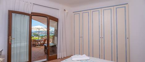 Ein Schlafzimmer mit einem Bett, einer Glastür zum Außenbereich mit Tisch und Stühlen sowie einem Kleiderschrank mit vertikalen Linien.