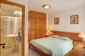 A bedroom with a wooden headboard, a single bed, a nightstand with a lamp, a wooden wardrobe, and a bathroom with a sink and mirror.