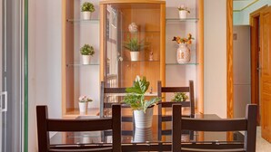 A dining area with a glass-top table, wooden chairs, and a wooden cabinet with shelves containing potted plants and decorative items.