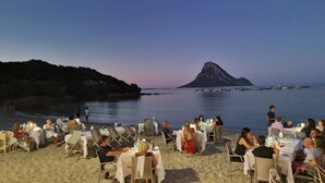 A beachfront restaurant with outdoor seating, tables, and chairs, overlooking the sea and a prominent hill. - Villa LUNA ROSSA with Sea View, Pool, A/C, Wi-Fi, Garden, Terrace & Balcony (san teodoro)