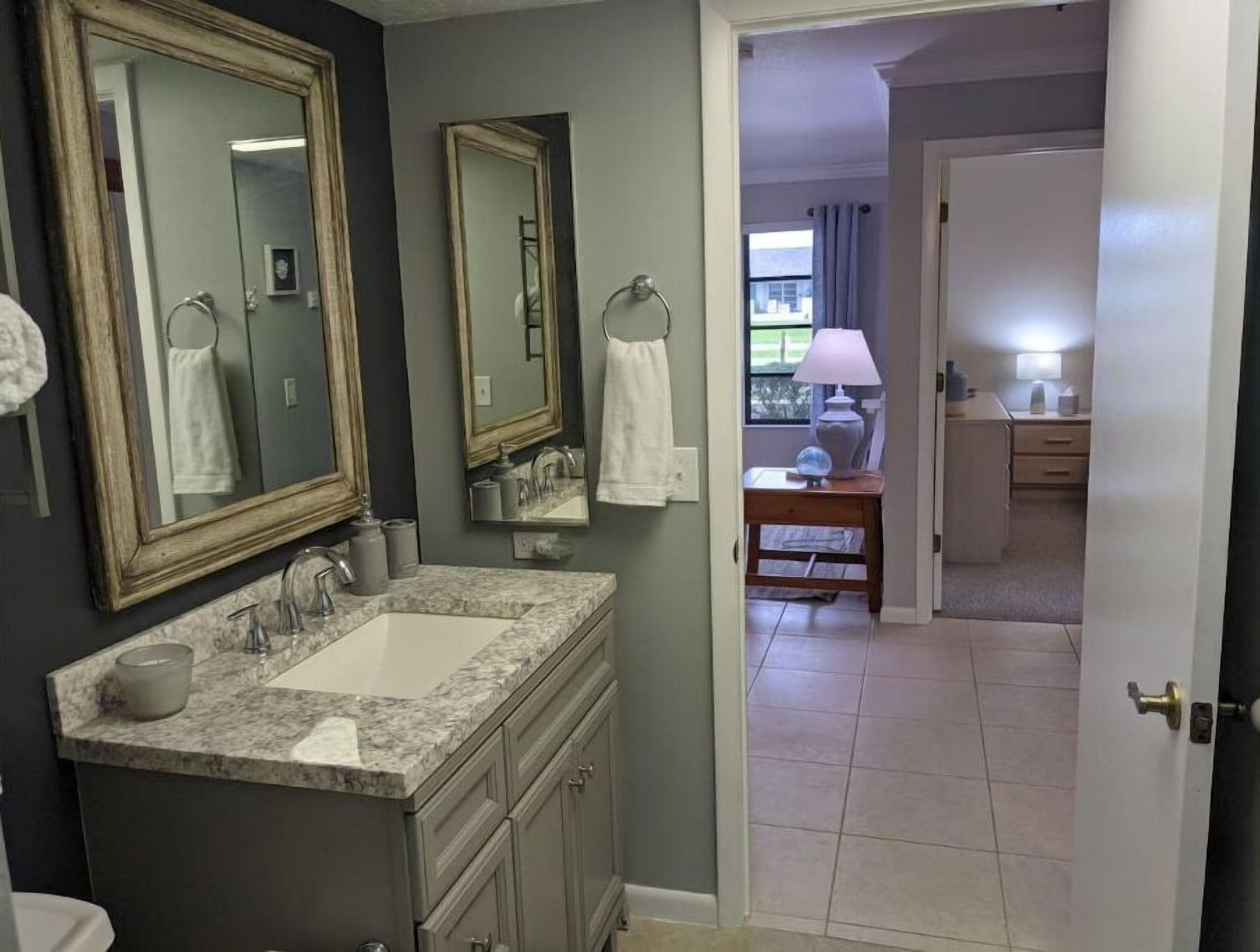 Une salle de bain avec un lavabo en marbre, deux miroirs et une vue sur une chambre à coucher équipée d’une table de chevet et d’une lampe.