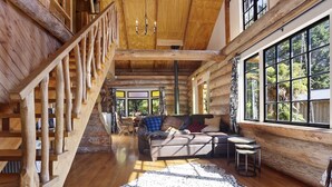 Interior d'una cabana espaiosa, amb una escala de fusta, una zona d'estar acollidora amb un sofà i cadires, i grans finestres que ofereixen vistes a l'exterior.