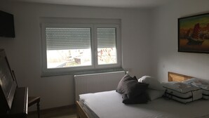 Un dormitorio con cama, almohadas, un radiador, una ventana con persianas, un cuadro en la pared y un escritorio con silla.