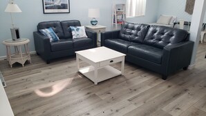 A living room with two black leather sofas, a white coffee table, and a wicker side table with a lamp. - The Deck House on the Silver Strand (Oxnard)