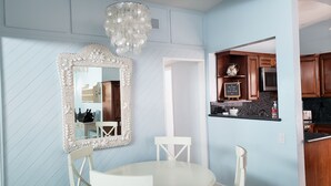 A dining area with a white round table, two white chairs, a decorative mirror, and a hanging light fixture. - The Deck House on the Silver Strand (Oxnard)