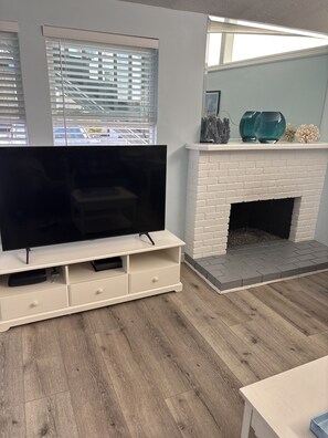 A living room with a flat-screen TV on a white entertainment unit, a white brick fireplace, and a window with blinds. - The Deck House on the Silver Strand (Oxnard)