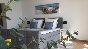 Un dormitorio con un sofá cama gris, un cuadro de una playa, una mesita de noche con lámpara y una alfombra estampada en el suelo.
