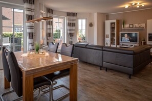 En moderne stue med sofa, spisebord med stole, fjernsyn og et stort vindue.