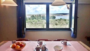 Un comedor con una mesa dispuesta para dos personas, un frutero y vistas a un lago y árboles a través de un ventanal grande.