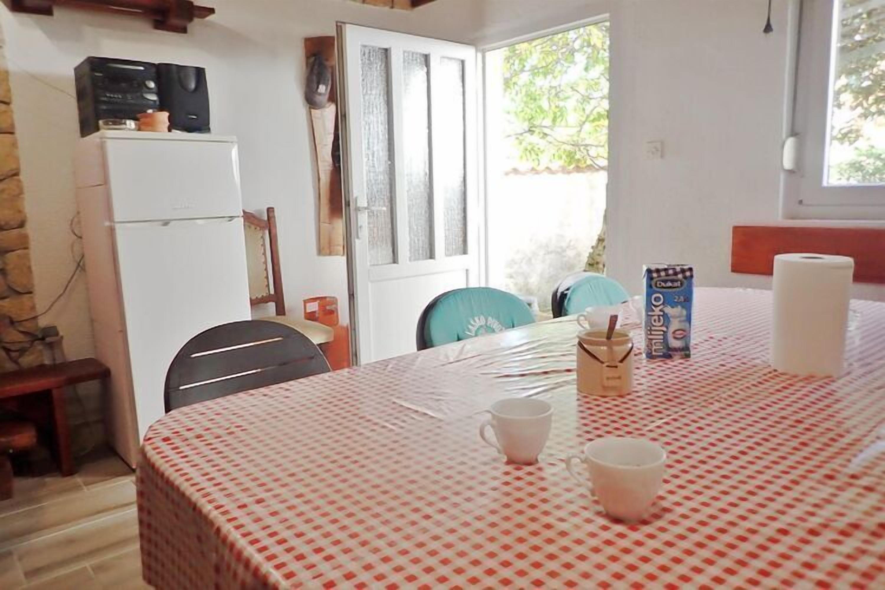 A small, cozy room with a bed, a table with a checkered tablecloth, a refrigerator, and a window with a view of greenery.