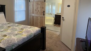 A bedroom with a bed, a ceiling fan, a window with blinds, a wooden wardrobe, a bathroom with a bathtub, and a television on a stand. - THE LIONS DEN  Home Next to  Lion & Tiger Reserve (Burlington)