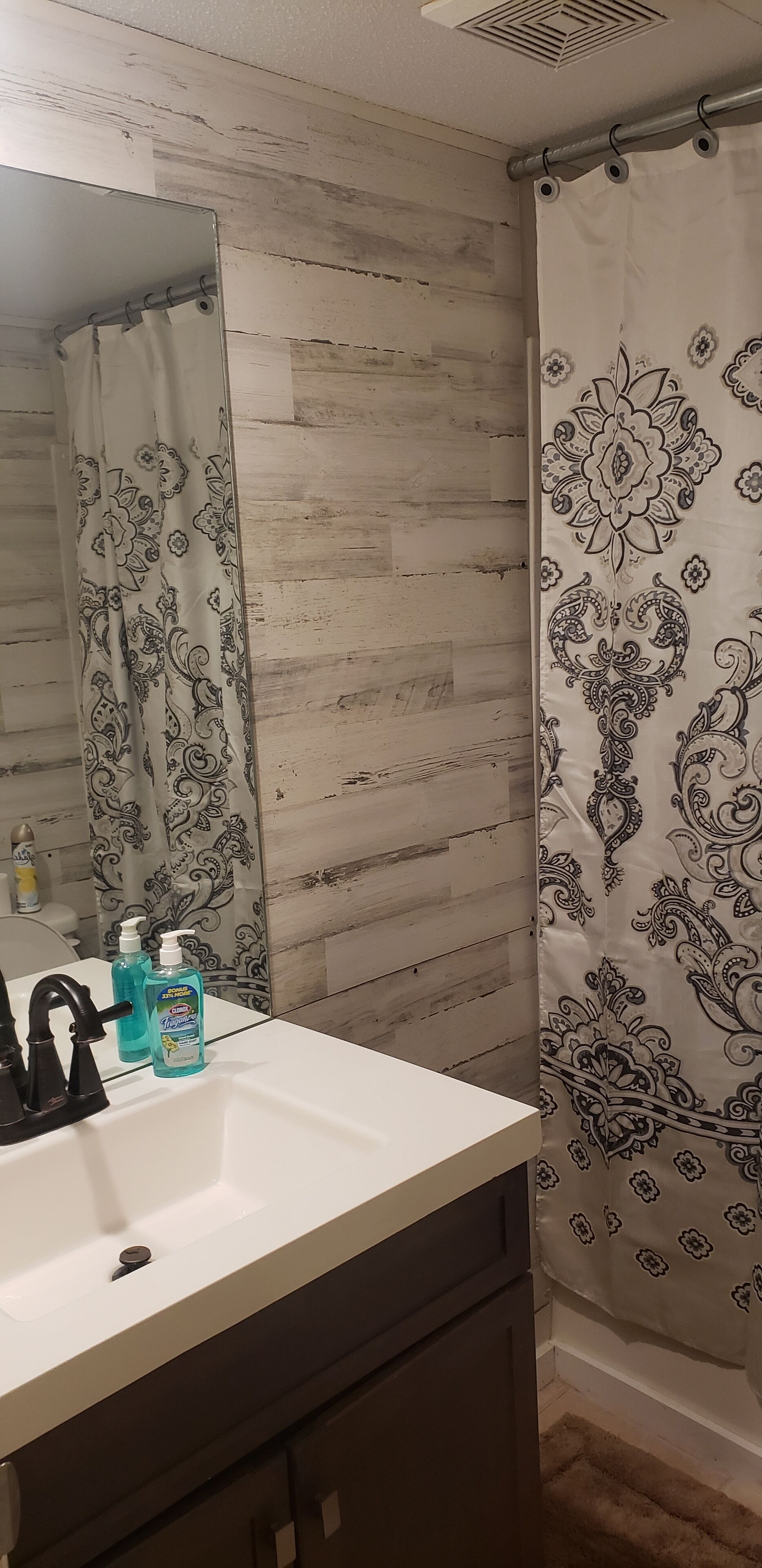 A bathroom with a wooden wall, a white sink with a black faucet, two bottles of blue liquid soap, and a shower curtain with a black floral pattern.