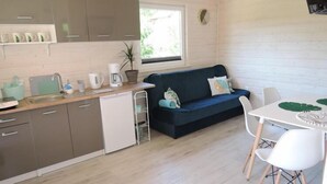 A compact kitchen with a sink, a coffee maker, and a plant. There is a dark blue sofa with cushions and a white table with chairs. - Bungalow in Kolczewo near Baltic Sea (Kolczewo)