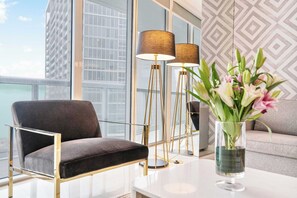 A modern living room with a dark upholstered chair, a white coffee table, and a vase with flowers. There are two floor lamps with brown shades and a patterned wall in the background.
