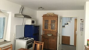 A compact kitchen with a stove, microwave, and blue refrigerator. There's a wooden cabinet with a bowl on top and a door leading to another room.