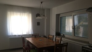 A dining area with a wooden table and chairs, a radiator, a window with blinds, and a kitchen area with a stove.