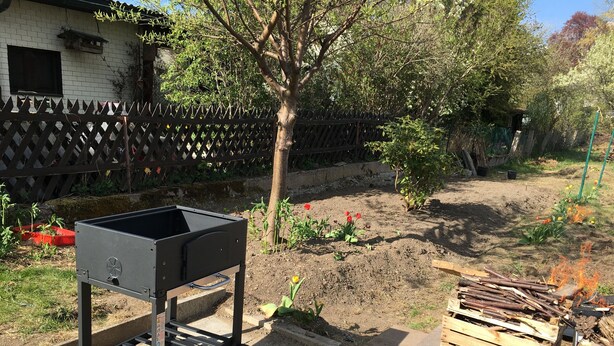 A barbecue grill in a backyard with a wooden fence, a tree, and a pile of firewood.