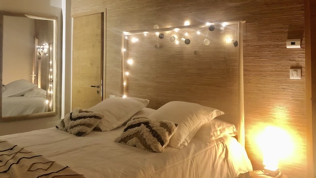 Une chambre avec une tête de lit en bois, un lit avec une literie blanche et des oreillers moelleux, un miroir et des guirlandes lumineuses.