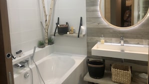 Une salle de bain moderne avec une baignoire, un lavabo et un miroir. On y trouve des articles de toilette sur une étagère et une petite plante.