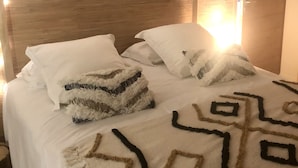 Un lit bien fait, avec du linge de lit blanc et des coussins décoratifs, une couverture à motifs et une guirlande lumineuse accrochée au chevet.