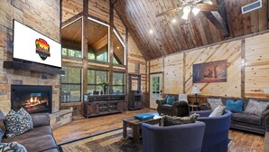 Ein geräumiges Blockhaus-Interieur mit Kamin aus Stein, Ledersofas und einem großen Fenster mit Blick auf einen Wald.