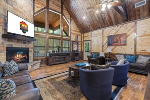 A spacious log cabin interior with a stone fireplace, leather sofas, and a large window overlooking a forest.