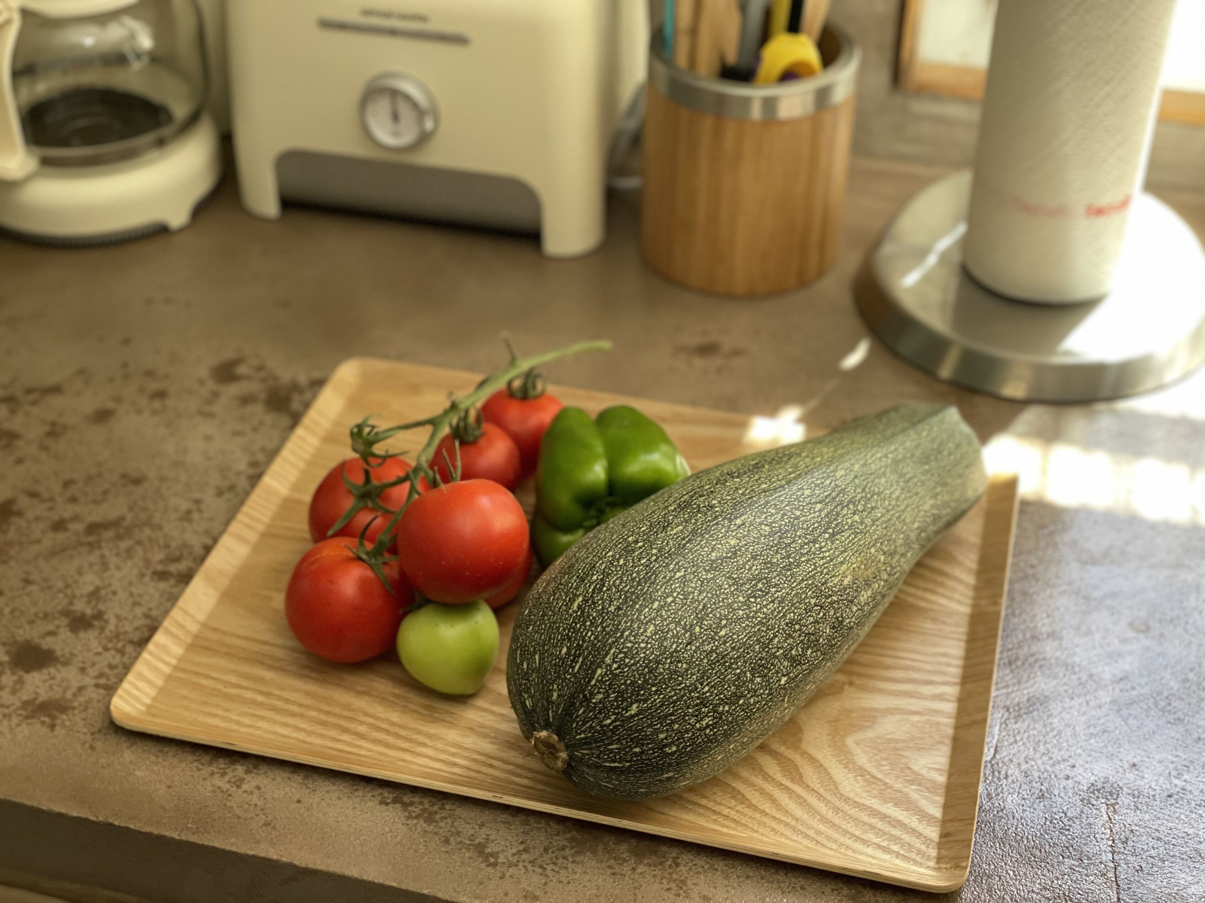 Puinen leikkuulauta, jonka päällä on suuri kesäkurpitsa, kirsikkatomaatteja ja vihreä paprika keittiön työtasolla.